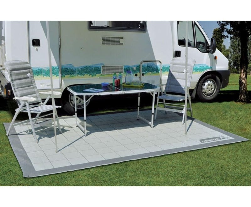 Tappeto da veranda per camper steso sull’erba sotto un tavolo e due sedie da campeggio.